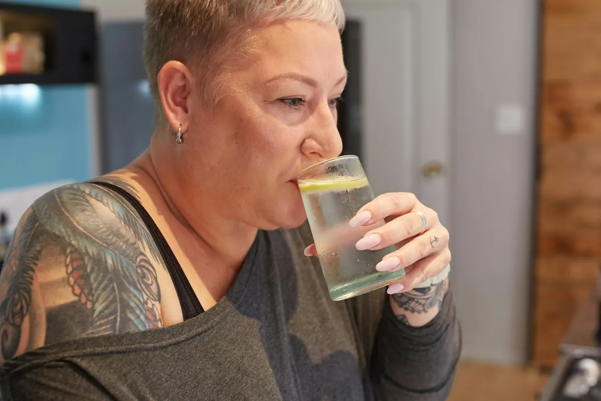 A woman with short silver hair drinks a glass of lemon water in her kitchen.
