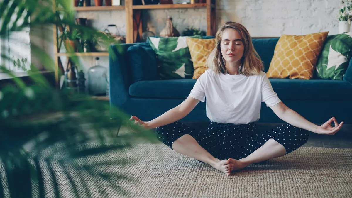 A woman sits cross-legged on the living room floor, eyes closed.